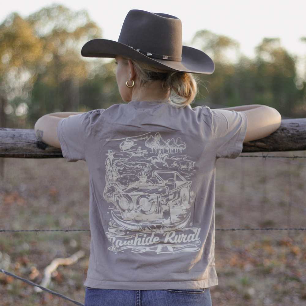 
                  
                    Woman wearing a Rawhide Rural t-shirt with a graphic design, standing by a wooden fence in a rural setting.
                  
                