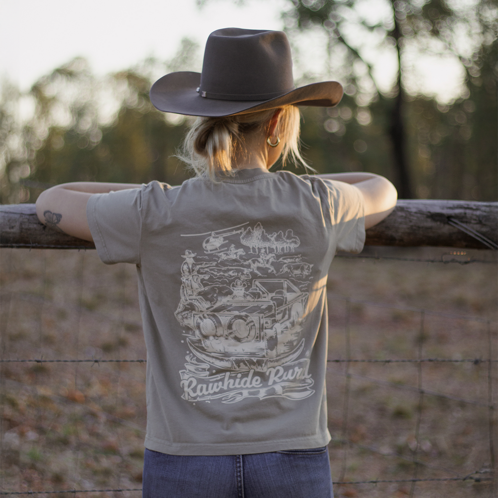 
                  
                    Woman wearing a Rawhide Rural t-shirt with a graphic design, standing by a fence with a natural background.
                  
                