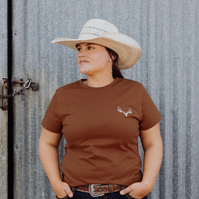 Woman standing in front of a grey background wearing a brown Rawhide Rural t-shirt.