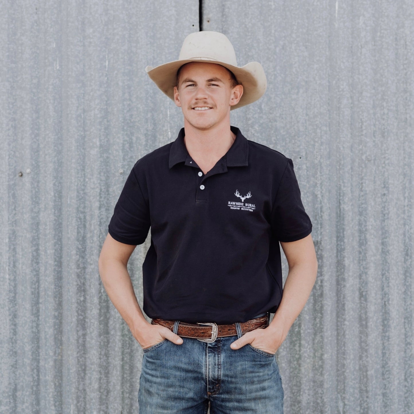 Man wearing a navy polo shirt with a logo and a cowboy hat, standing against a corrugated metal wall.