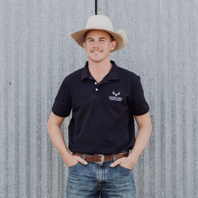 Man wearing a navy polo shirt with a logo and a cowboy hat, standing against a corrugated metal wall.