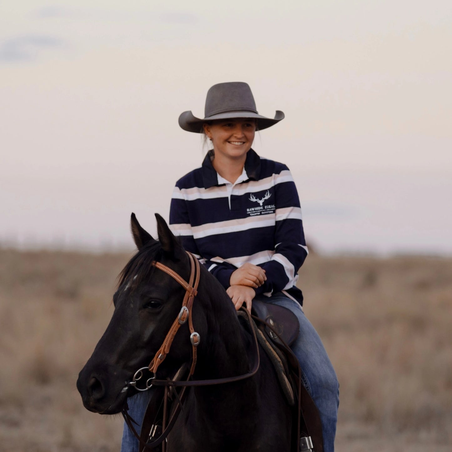 Person riding a horse in an open field with a neutral background wearing a navy, pink and white rugby jumper.