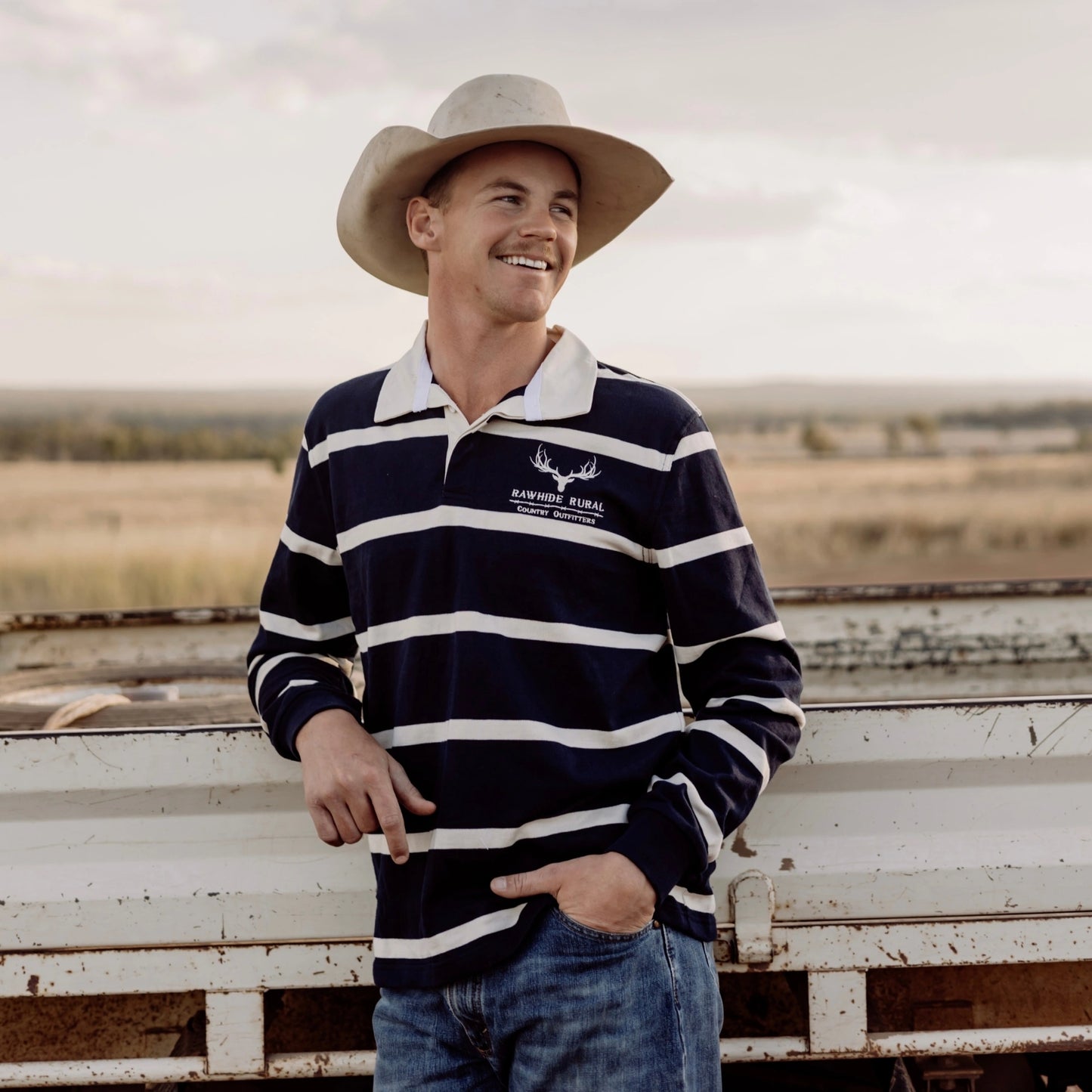 Man wearing a striped shirt and cowboy hat standing next to a ute tray with a rural landscape in the background