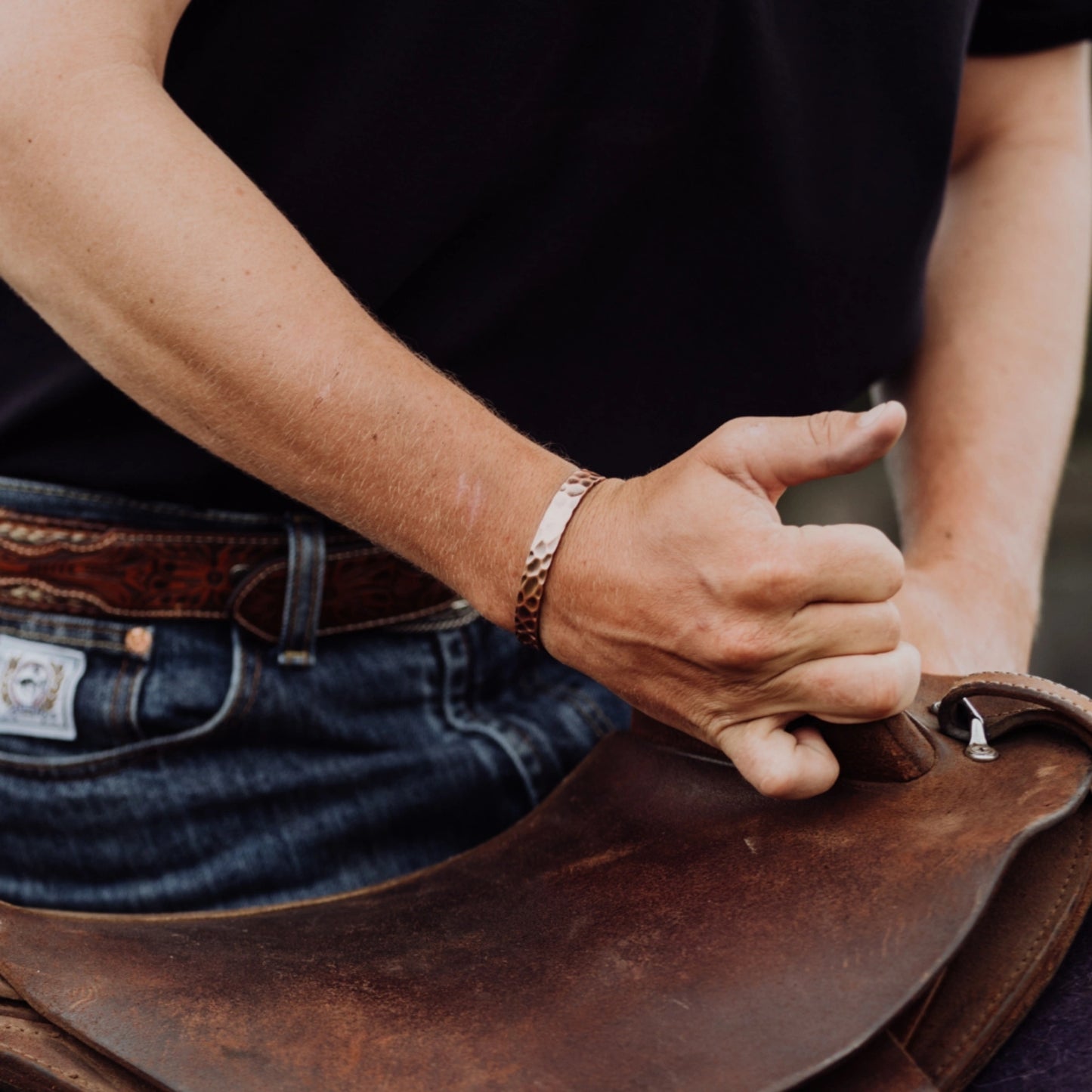 Person adjusting a brown leather saddle with a focus on the hands and saddle, wearing a dark beaten copper band.