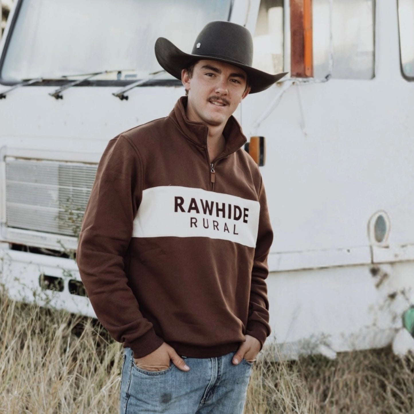 Man wearing a brown 'Rawhide Rural' sweatshirt in front of a vintage truck.