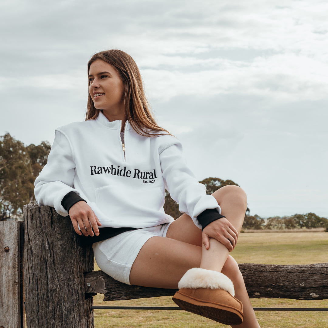 Woman wearing a white 'Rawhide Rural' jumper sitting on a wooden fence outdoors.