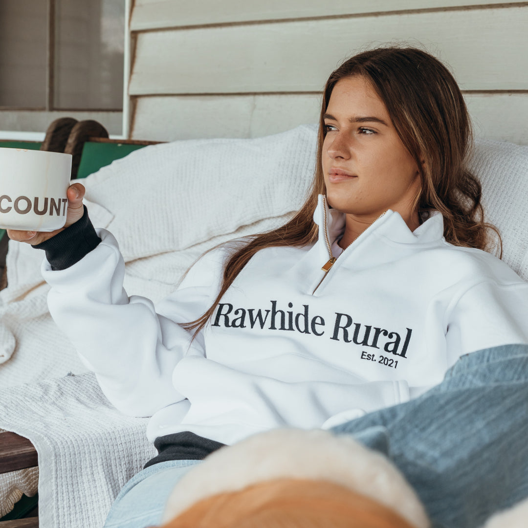Woman wearing a 'Rawhide Rural' jumper, holding a mug, sitting on an outdoor chair.