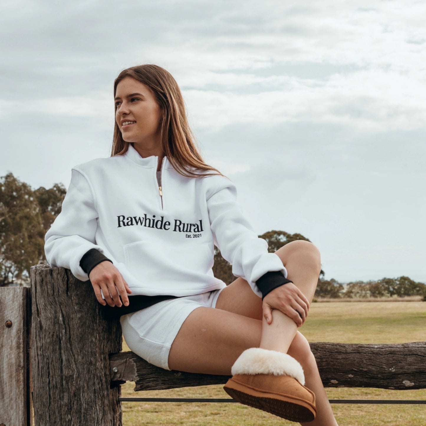 Woman wearing a white 'Rawhide Rural' sweatshirt sitting on a wooden post outdoors.