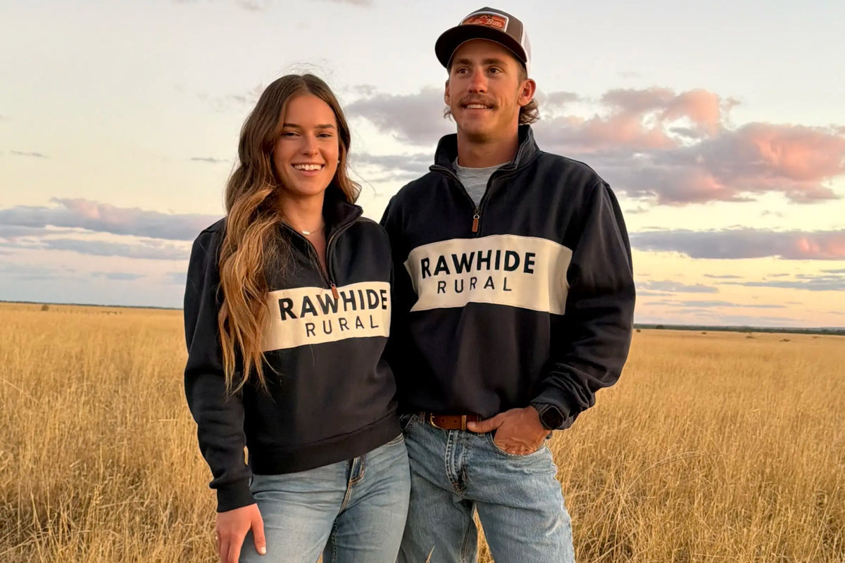 A man and a woman standing in a dry grass field at sunset, both wearing matching navy quarter zip jumper with a white horizontal stripe across the chest that reads 'RAWHIDE RURAL'.