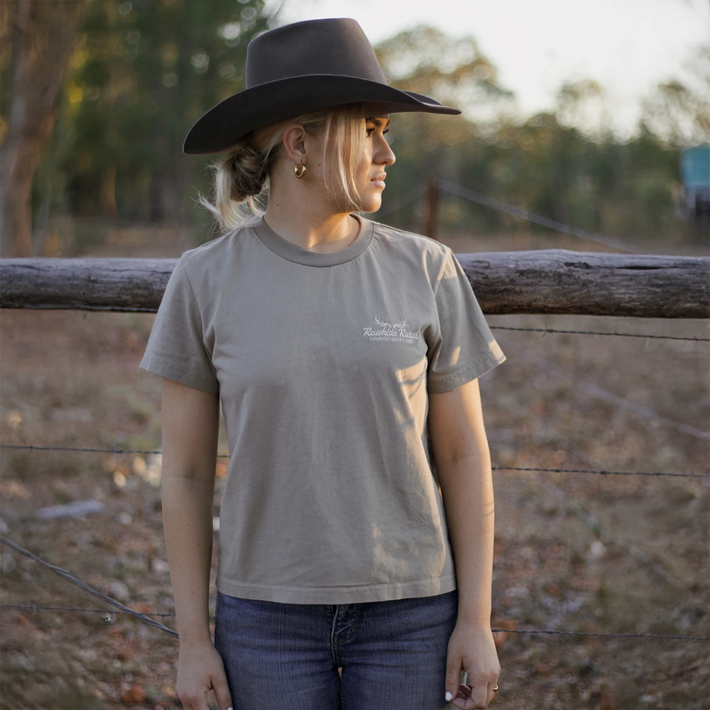 
                  
                    Woman wearing a Rawhide Rural t-shirt and cowboy hat standing near a wooden fence with trees in the background.
                  
                