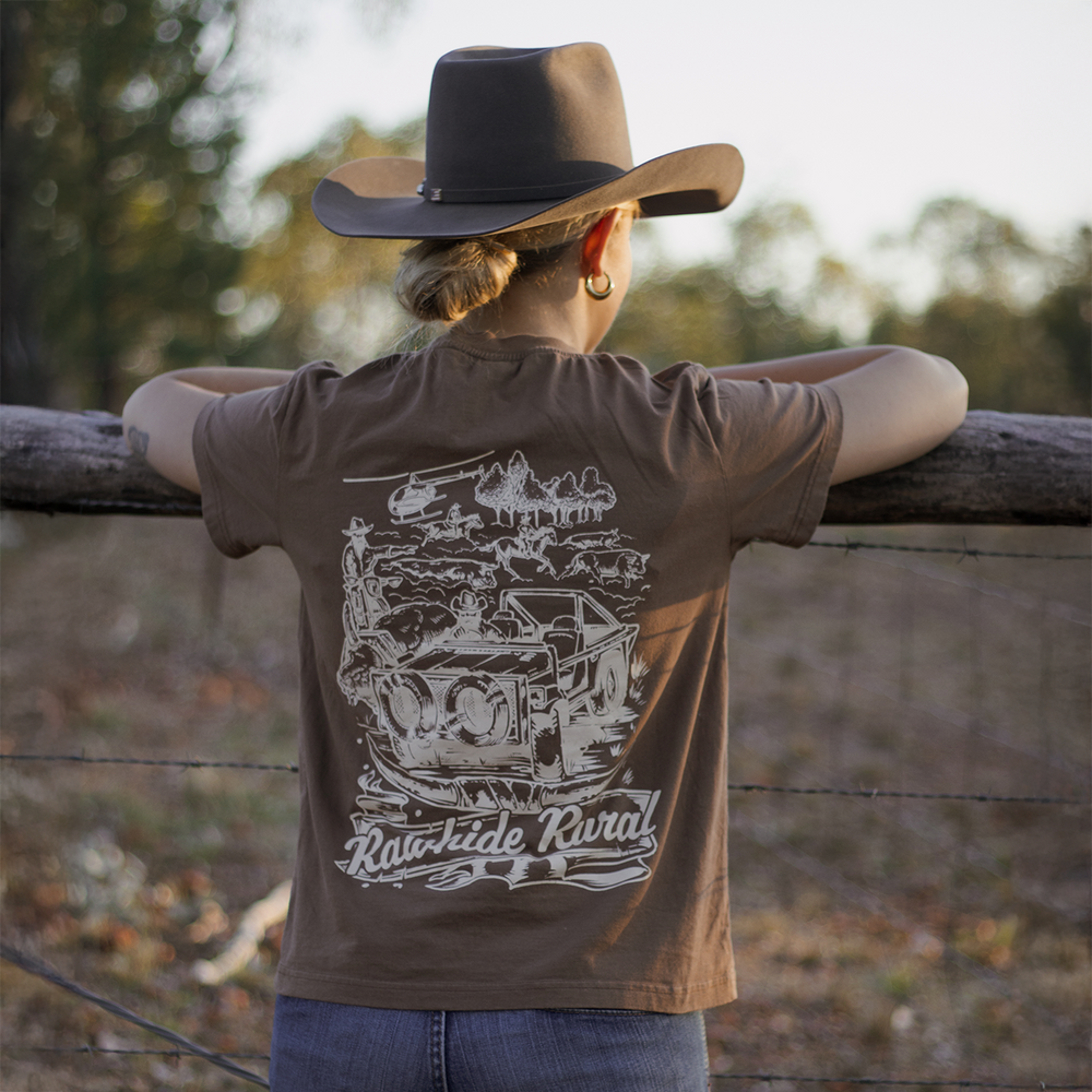 
                  
                    Woman wearing a Rawhide Rural brown shirt with a graphic design, standing by a wooden fence in a natural setting.
                  
                