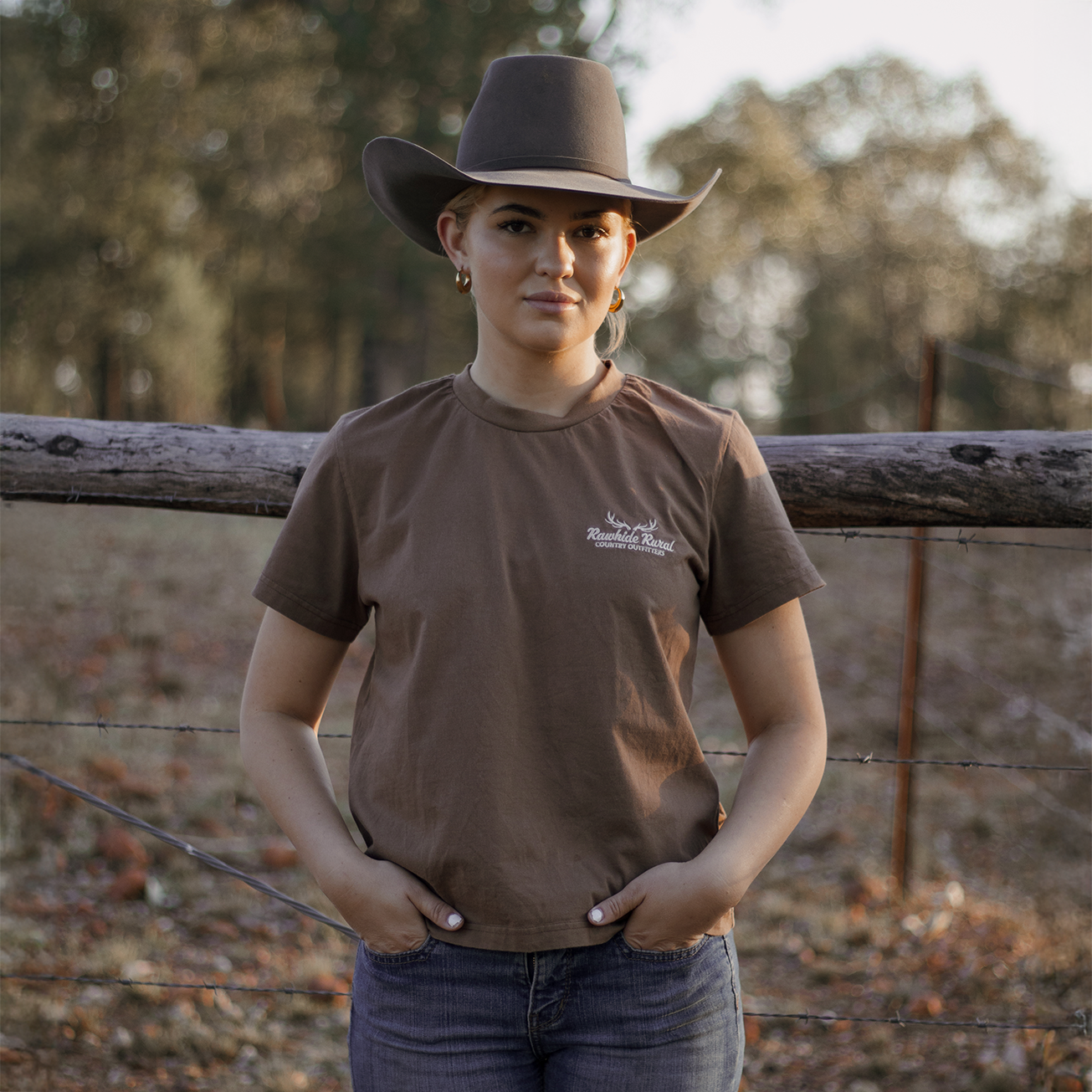 Woman wearing a brown hat and brown Rawhide Rural t-shirt standing in front of a wooden fence with trees in the background.