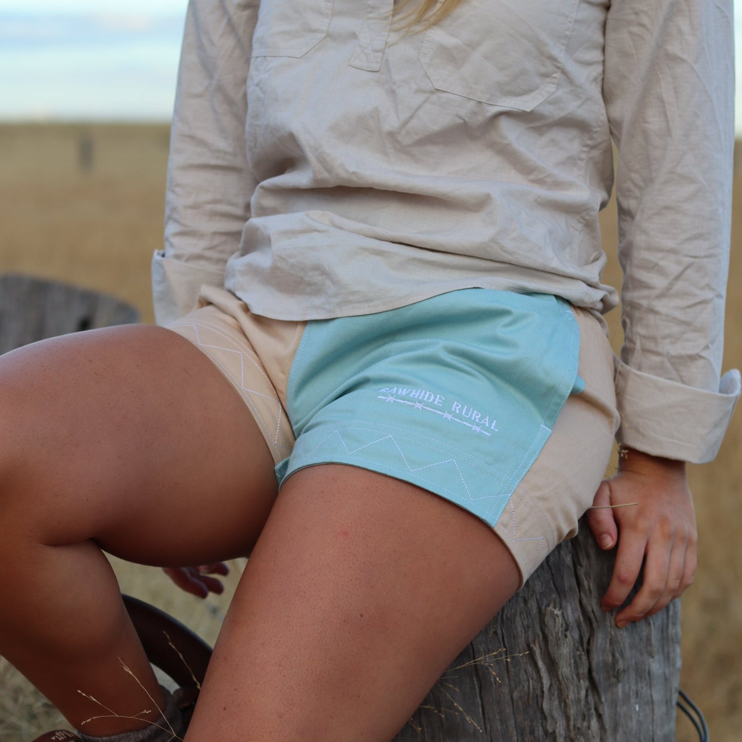 Person wearing a beige shirt and light blue shorts sitting on a wooden post with a natural background.